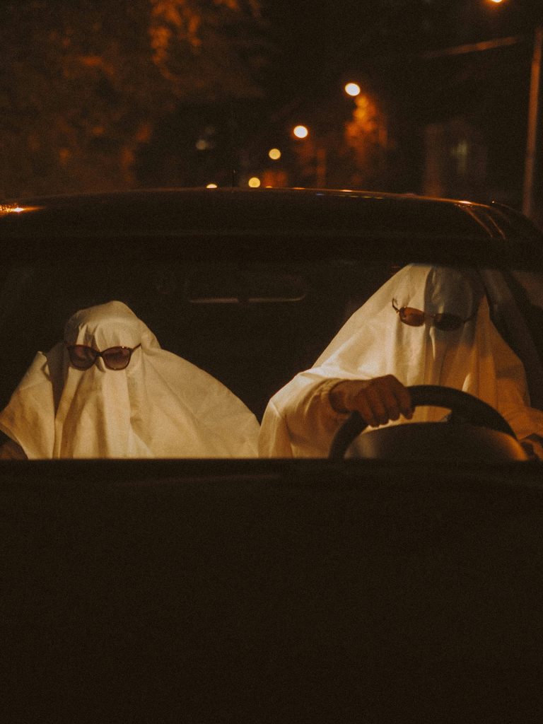 Two people dressed as ghosts with white sheets and sunglasses sit in a car at night, with the driver holding the steering wheel. The scene is dimly lit, giving a spooky Halloween vibe.