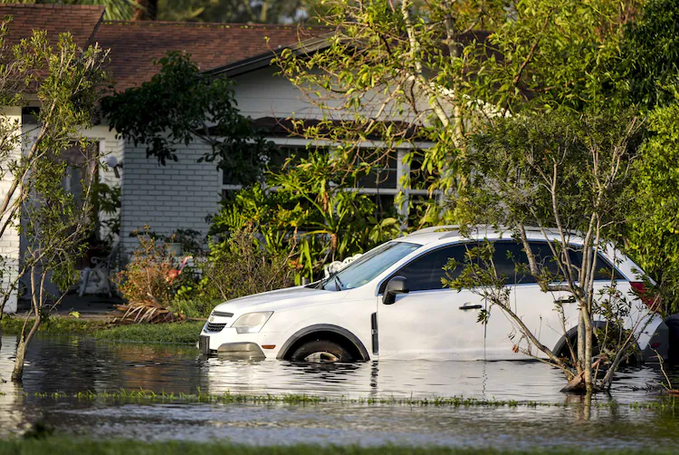 A white SUV partially submerged in floodwater near a residential house, surrounded by green trees and plants, likely due to hurricane flooding.
