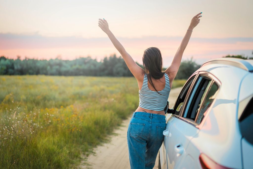 : A woman in a striped tank top and jeans stands with her arms raised, enjoying the sunset in a scenic field next to a white car.