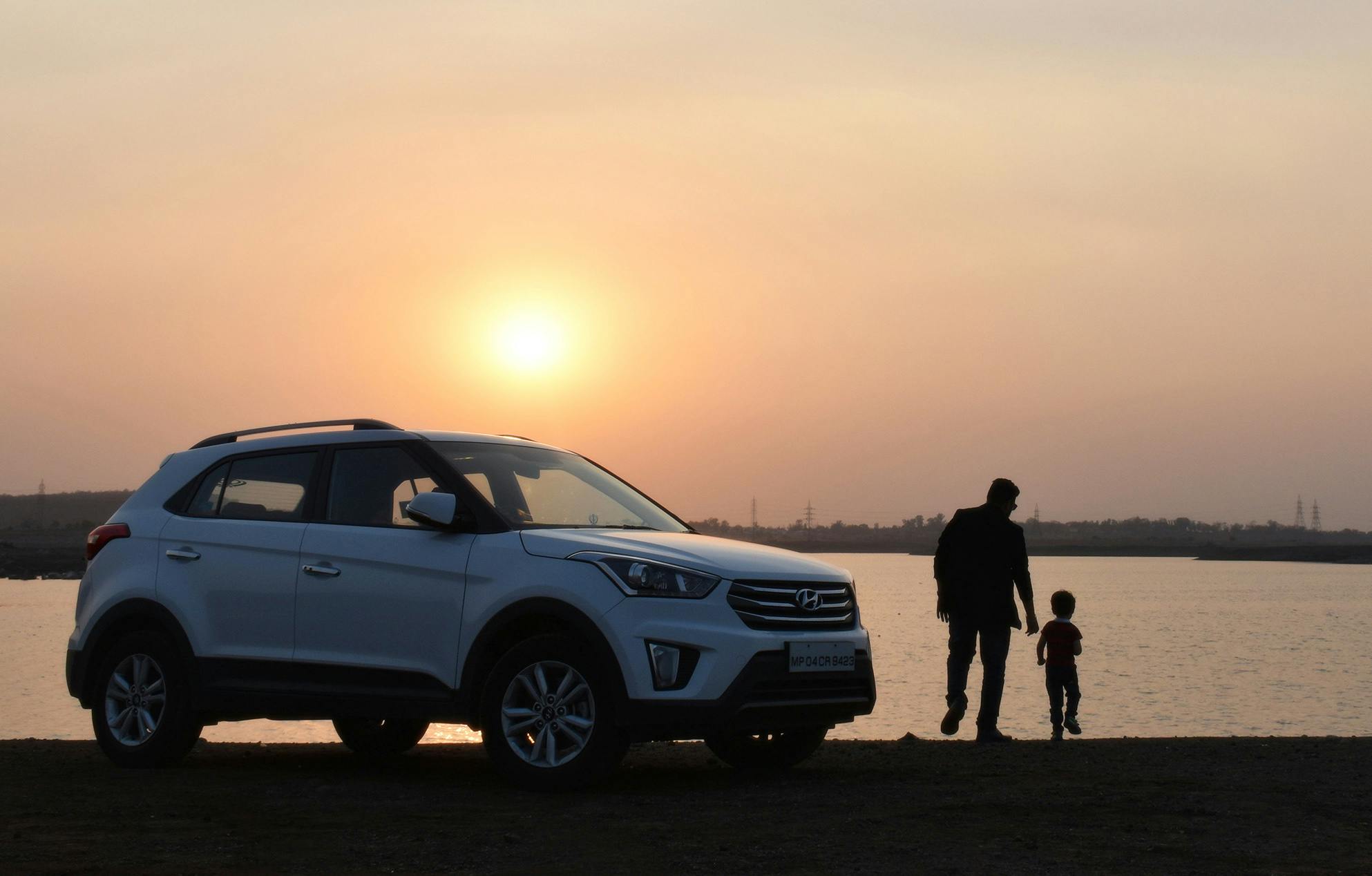 White SUV parked near a lake at sunset with a man and child walking together in the background.